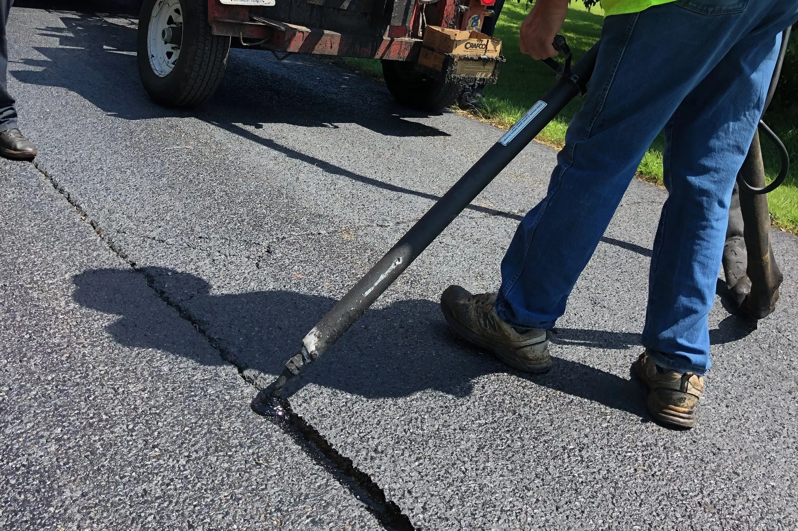 Worker applying hot rubberized sealant into a pavement crack with a long-handled wand, the sealant melter trailer behind