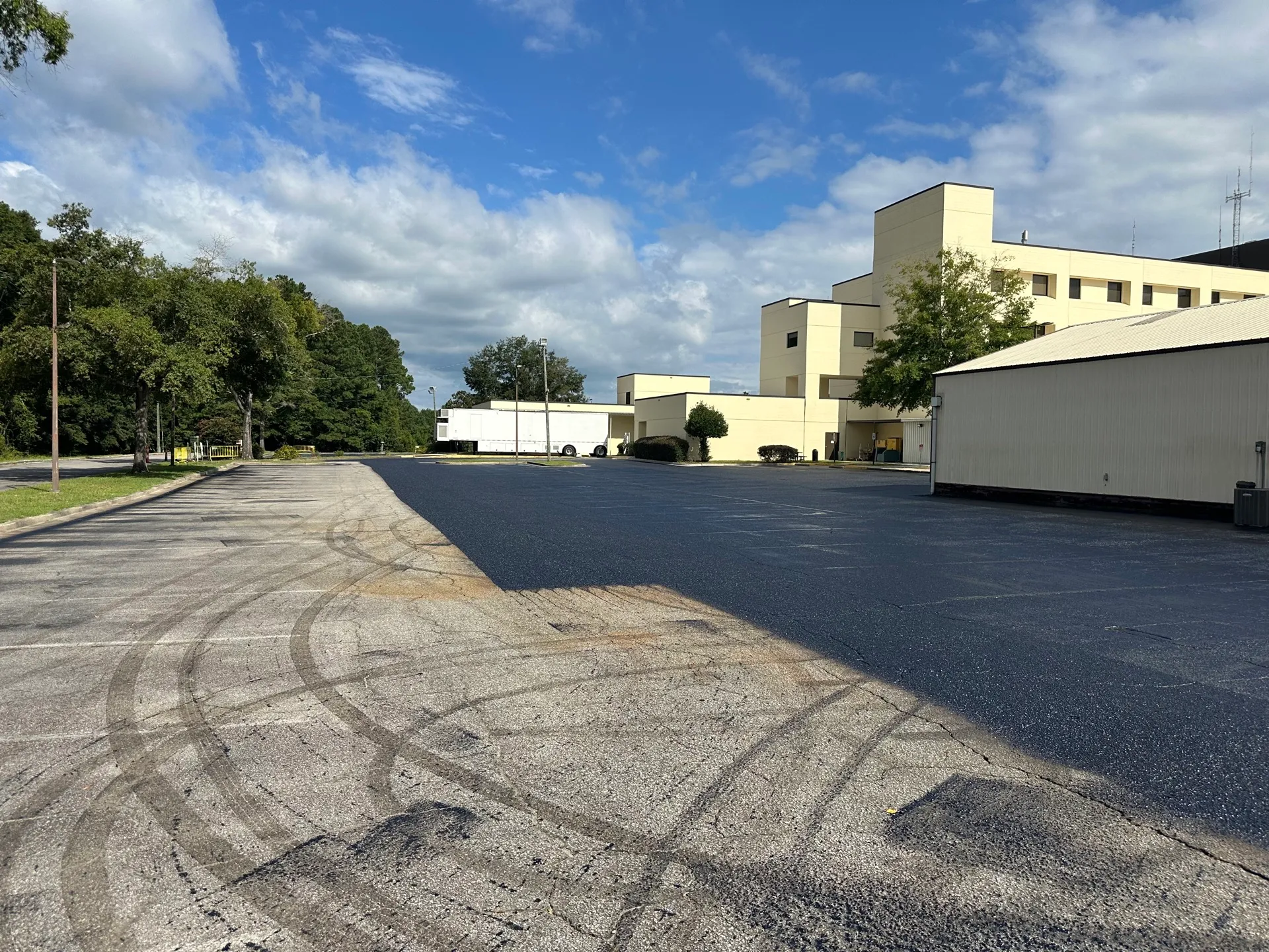 Freshly sealed industrial lot meeting older pavement, with distribution buildings and a trailer beyond.