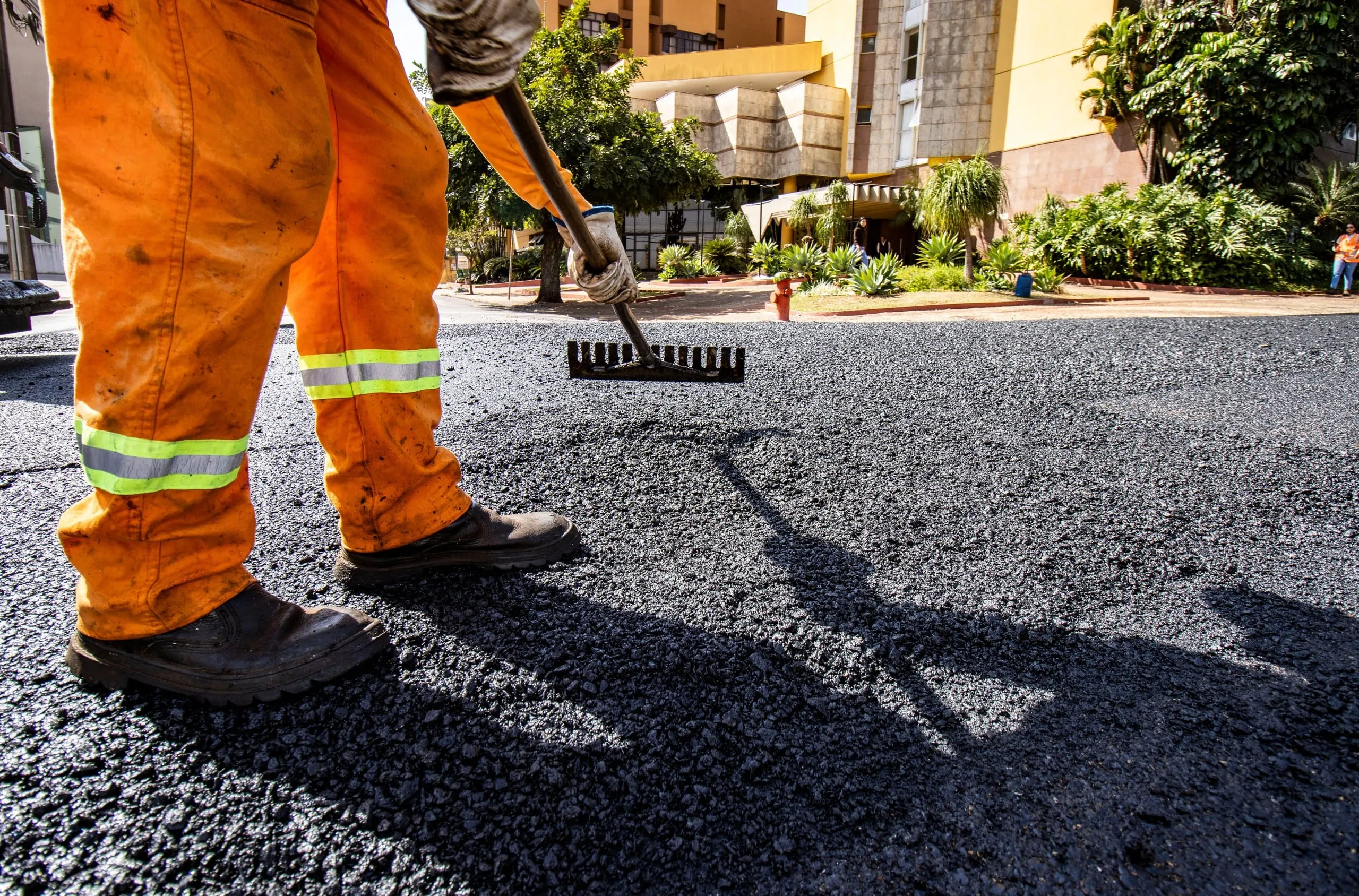 Worker in orange hi-vis raking out a fresh layer of hot black asphalt on a city street