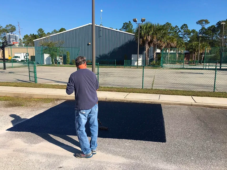Applicator spreading a dark rejuvenator sample square on faded asphalt beside a fenced recreation court