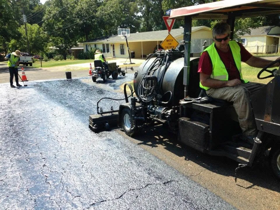 TriTechnologies spray units applying asphalt rejuvenator across a neighborhood street, with founder Jeff Pokorny at the controls and a crew member brooming the treated surface.