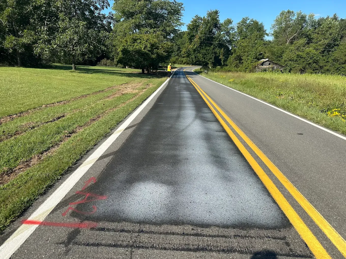 A rural two-lane road freshly restored with plant-based rejuvenator — dark, renewed asphalt with crisp double-yellow centerline.